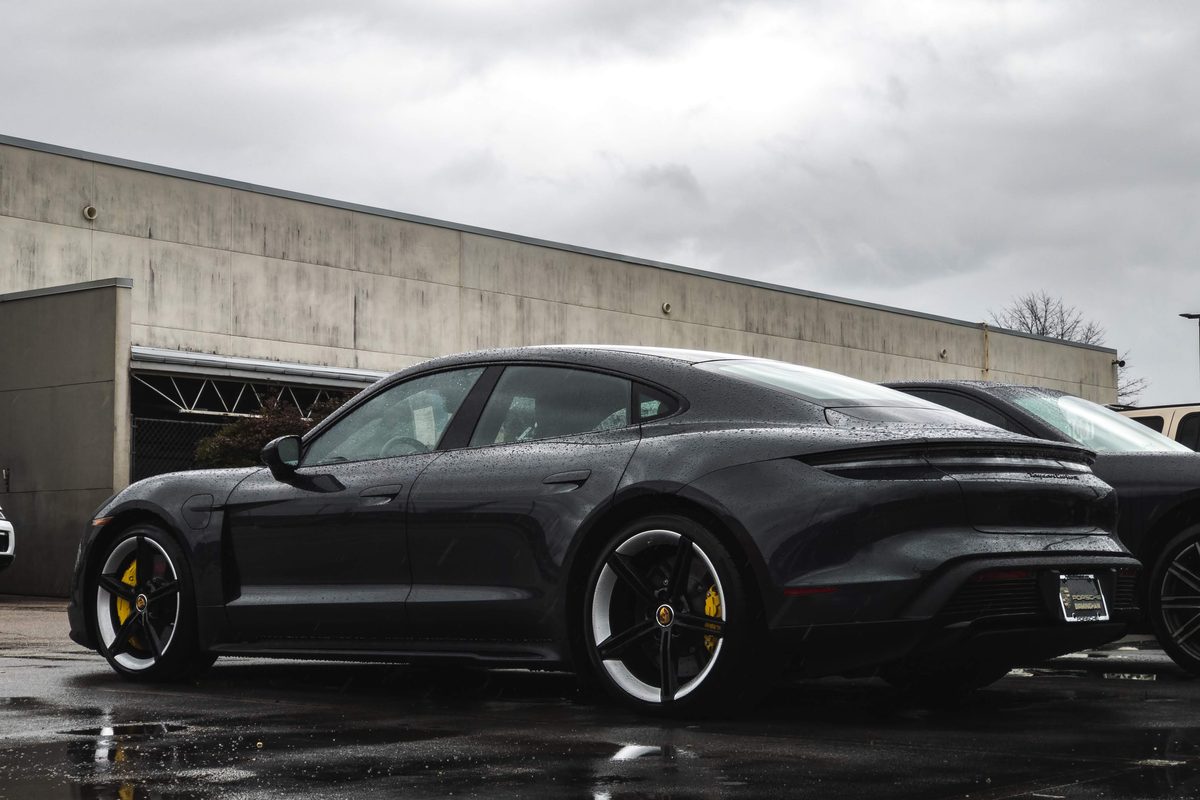 A dark grey Porsche Taycan parked on wet asphalt outside a concrete building under heavy overcast.