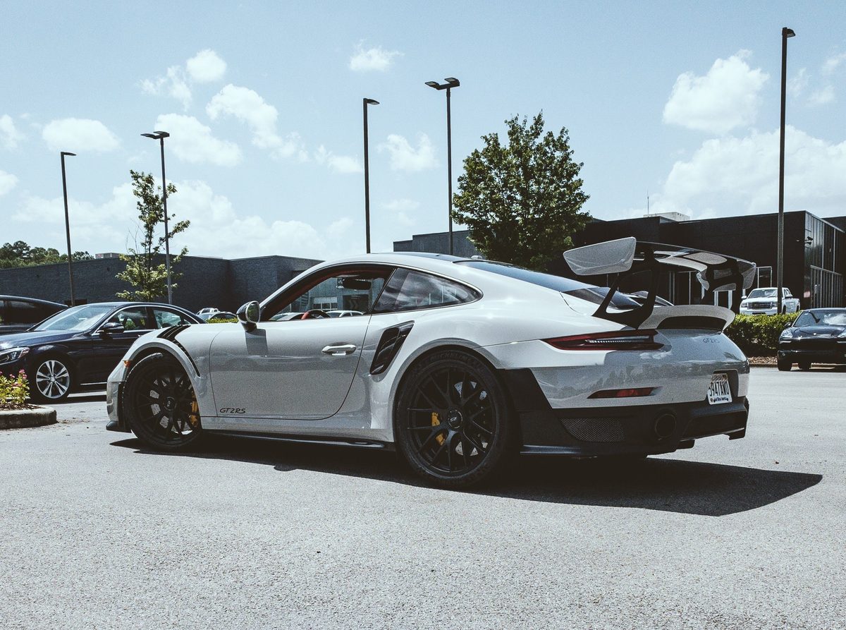 A white Porsche 911 GT2 RS with its rear wing raised, parked in a dealership lot under summer cloud.