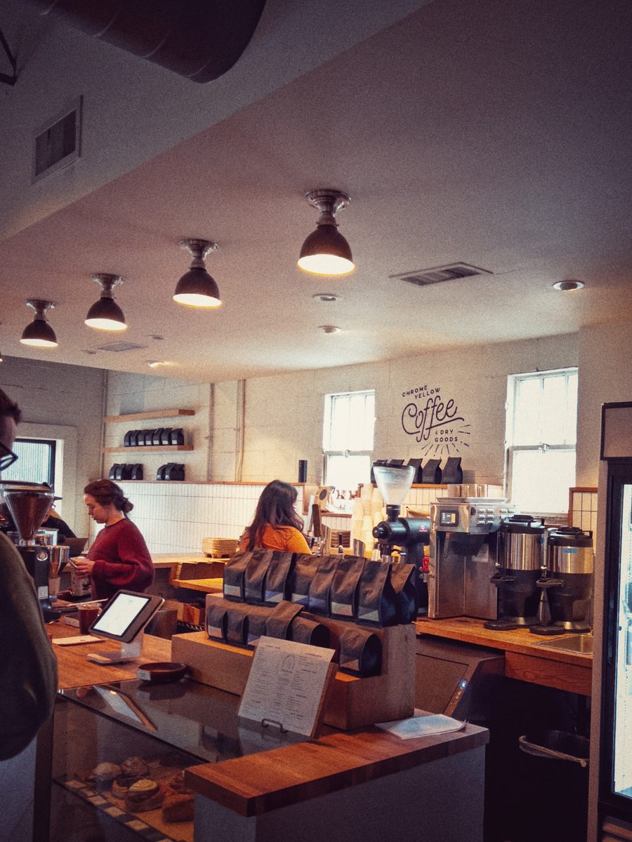The counter of a coffee shop, two staff working behind rows of coffee bags under pendant lamps.