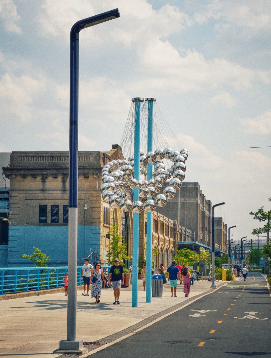A waterfront promenade on a summer afternoon, people walking past a suspended cluster sculpture beside a bike lane.