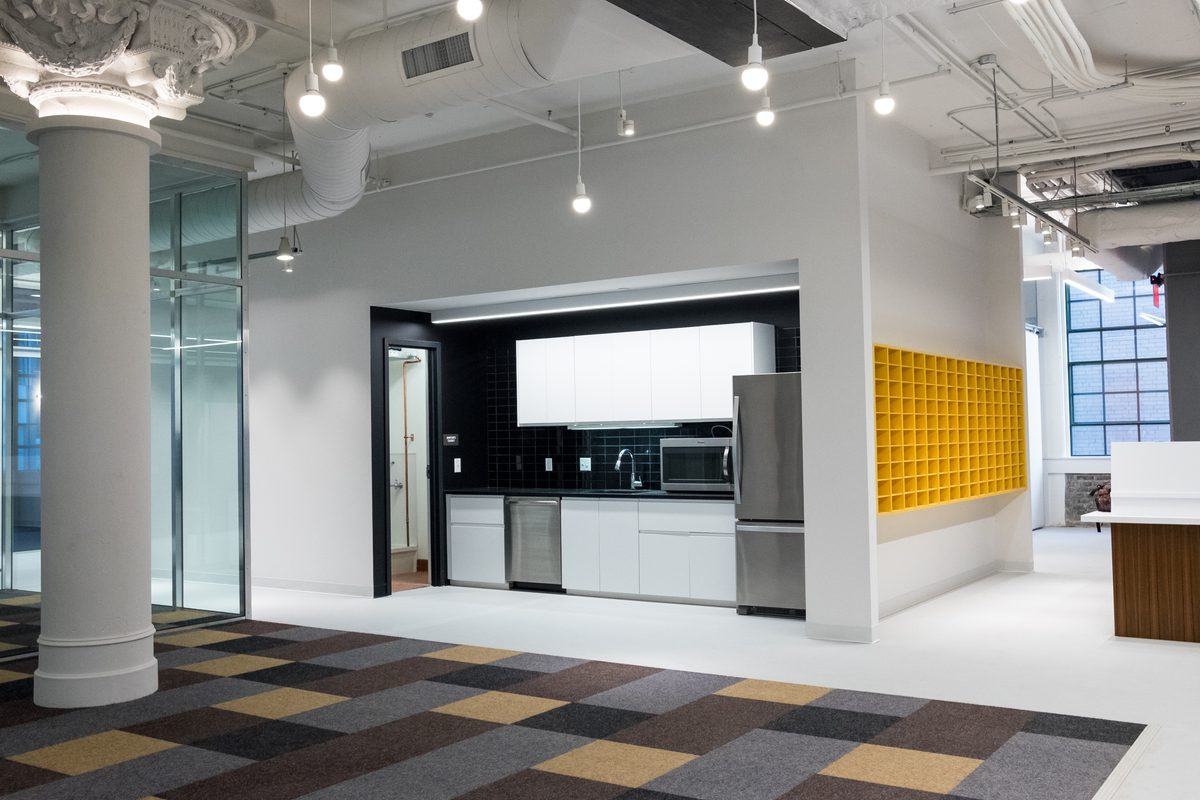An open-plan office interior with a kitchenette, exposed ceiling ducts and a yellow tiled wall.
