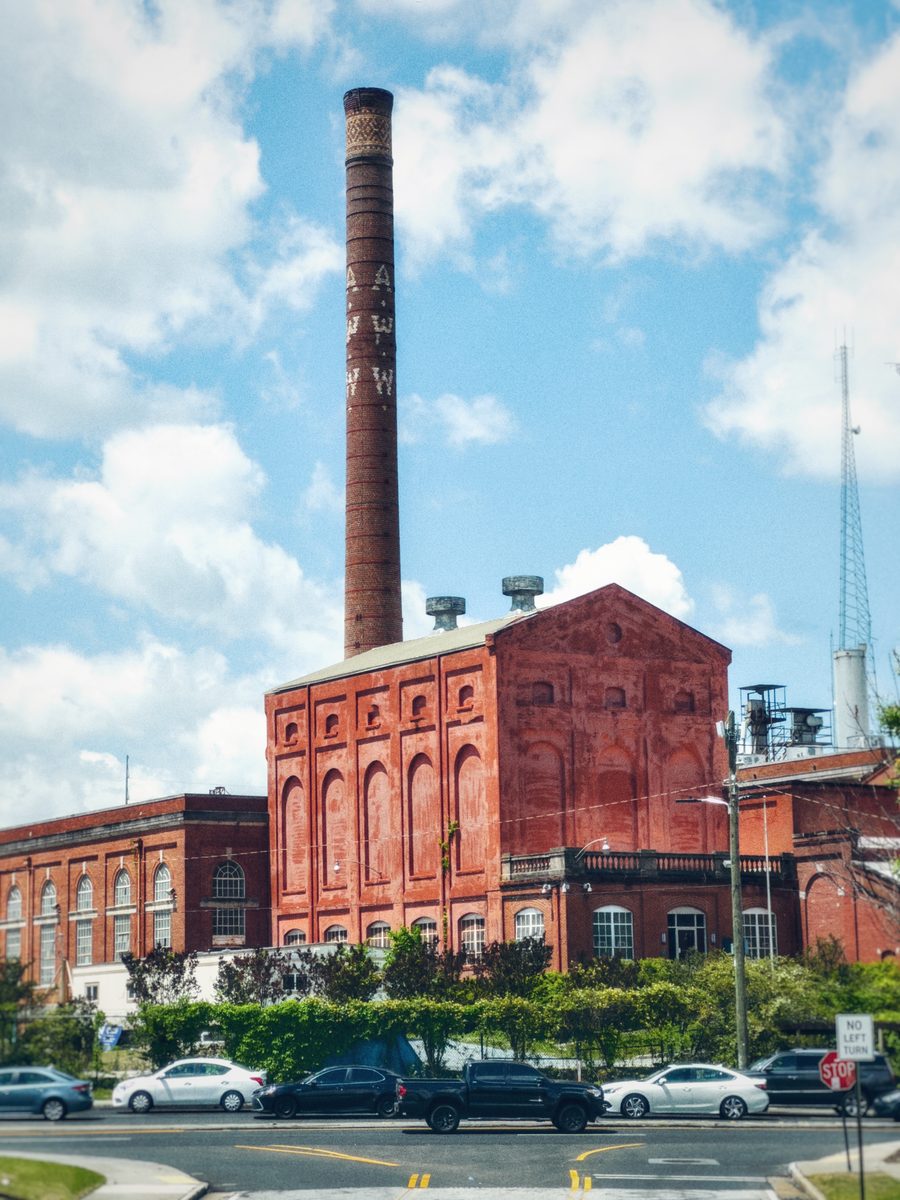 A red brick industrial building and its tall lettered smokestack against summer cloud, traffic passing in front.
