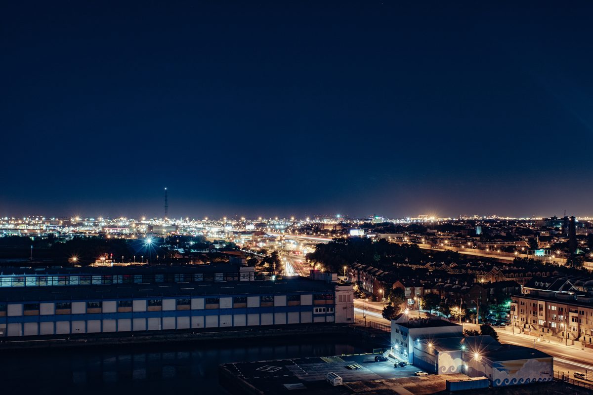 A port district at night seen from a rooftop, streets and warehouses lit against a deep blue sky.