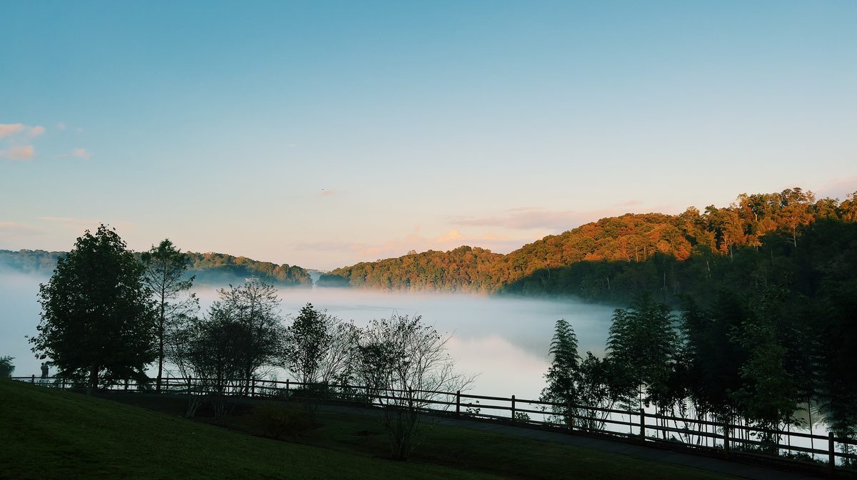 Mist lying on a reservoir at first light, wooded hills catching the sun behind a rail fence.