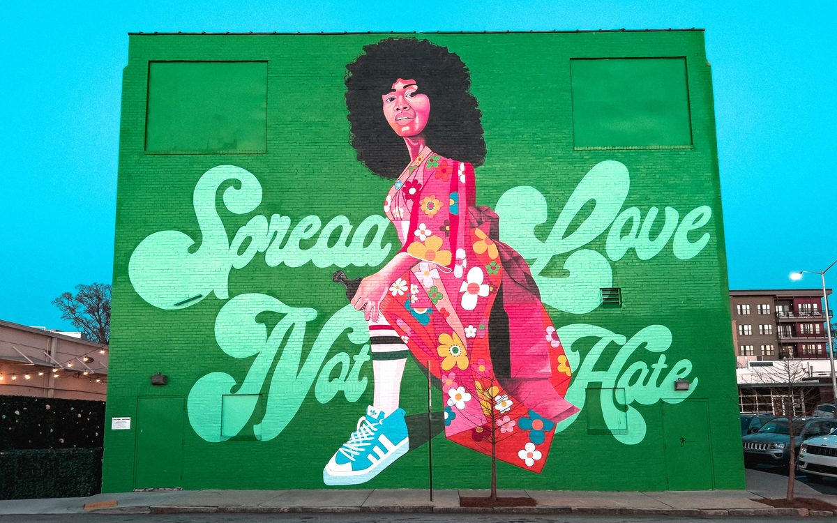 A green painted wall mural reading Spread Love Not Hate, a woman in a floral dress stepping across it, at dusk.
