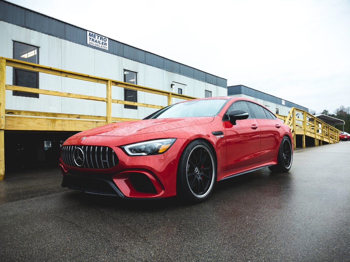 A red Mercedes-AMG GT 63 on wet ground in front of a yellow-railed warehouse, under flat overcast.