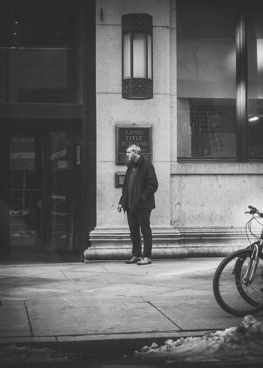 A man standing alone outside the Land Title Building in winter, a bicycle at the frame edge, in black and white.