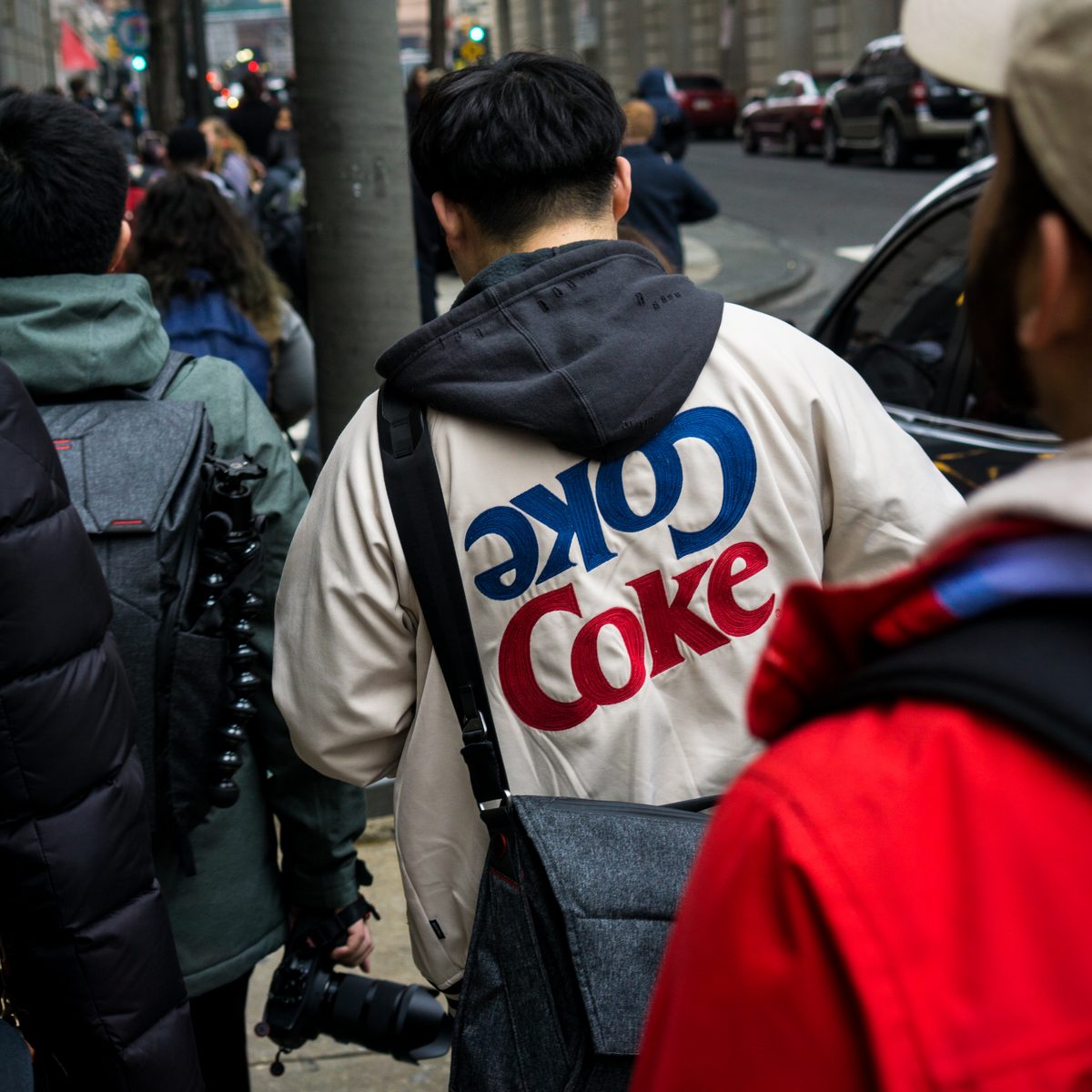 A crowded pavement seen from behind, a photographer in a Coke jacket carrying a camera among the shoulders.