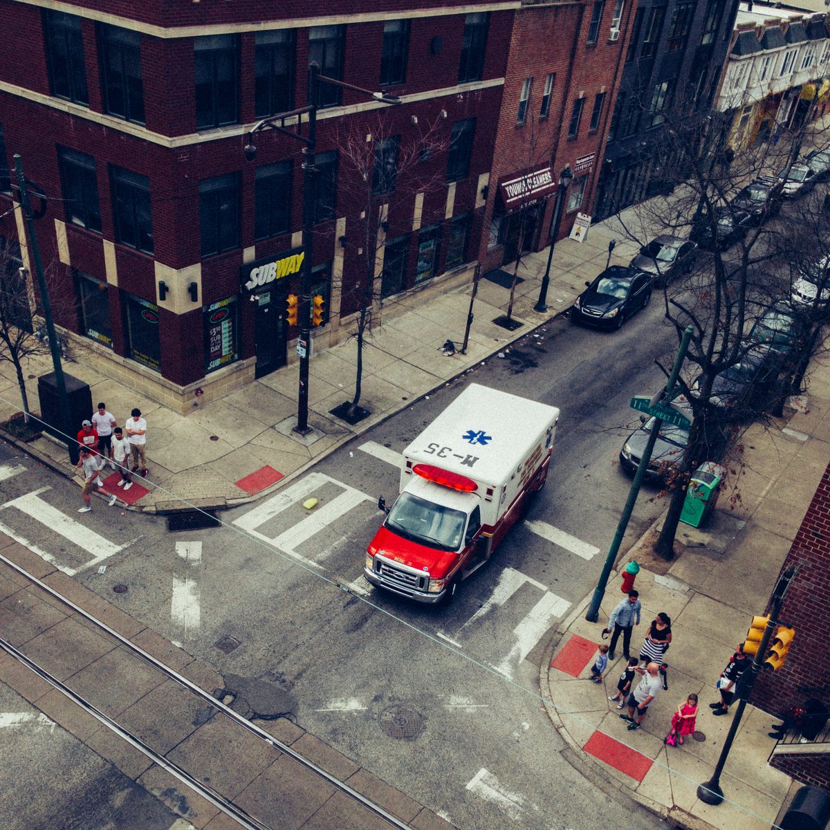 An ambulance crossing a wet intersection, seen looking down from above, pedestrians waiting on the corners.