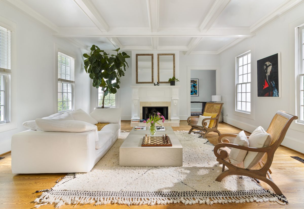 A white living room with a coffered ceiling, a chess set on the table and cane chairs by the fireplace.
