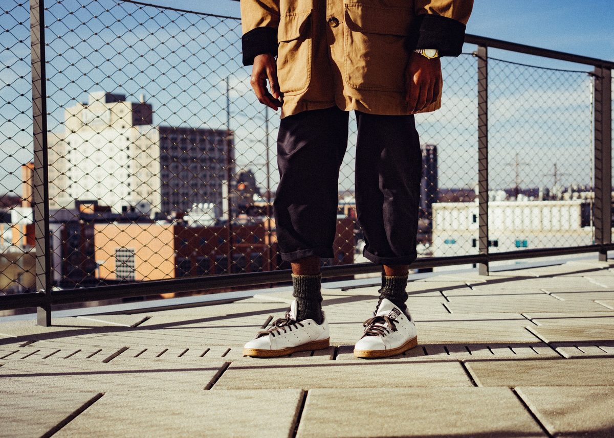 A figure from the waist down on a rooftop in hard sun, city rooftops through the chain-link fence behind.