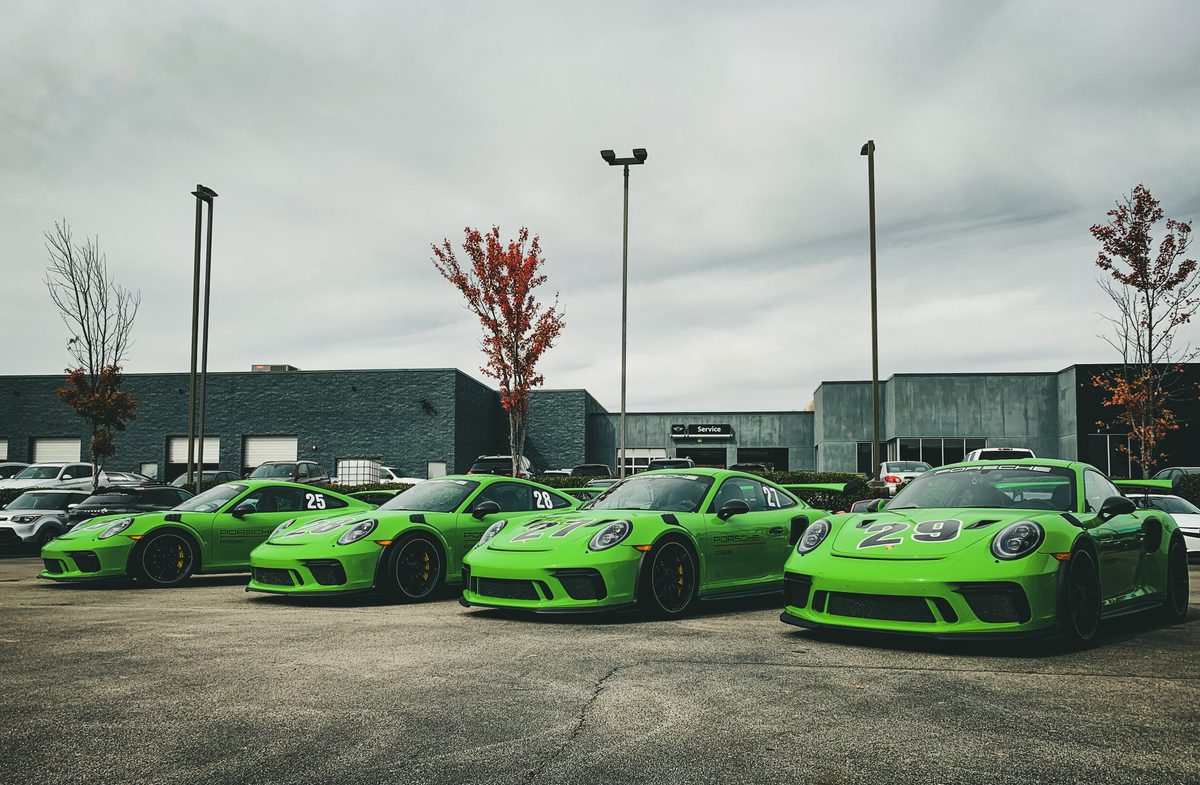 Four green Porsche 911 GT3 RS lined up nose-out in a lot, autumn trees behind under grey sky.