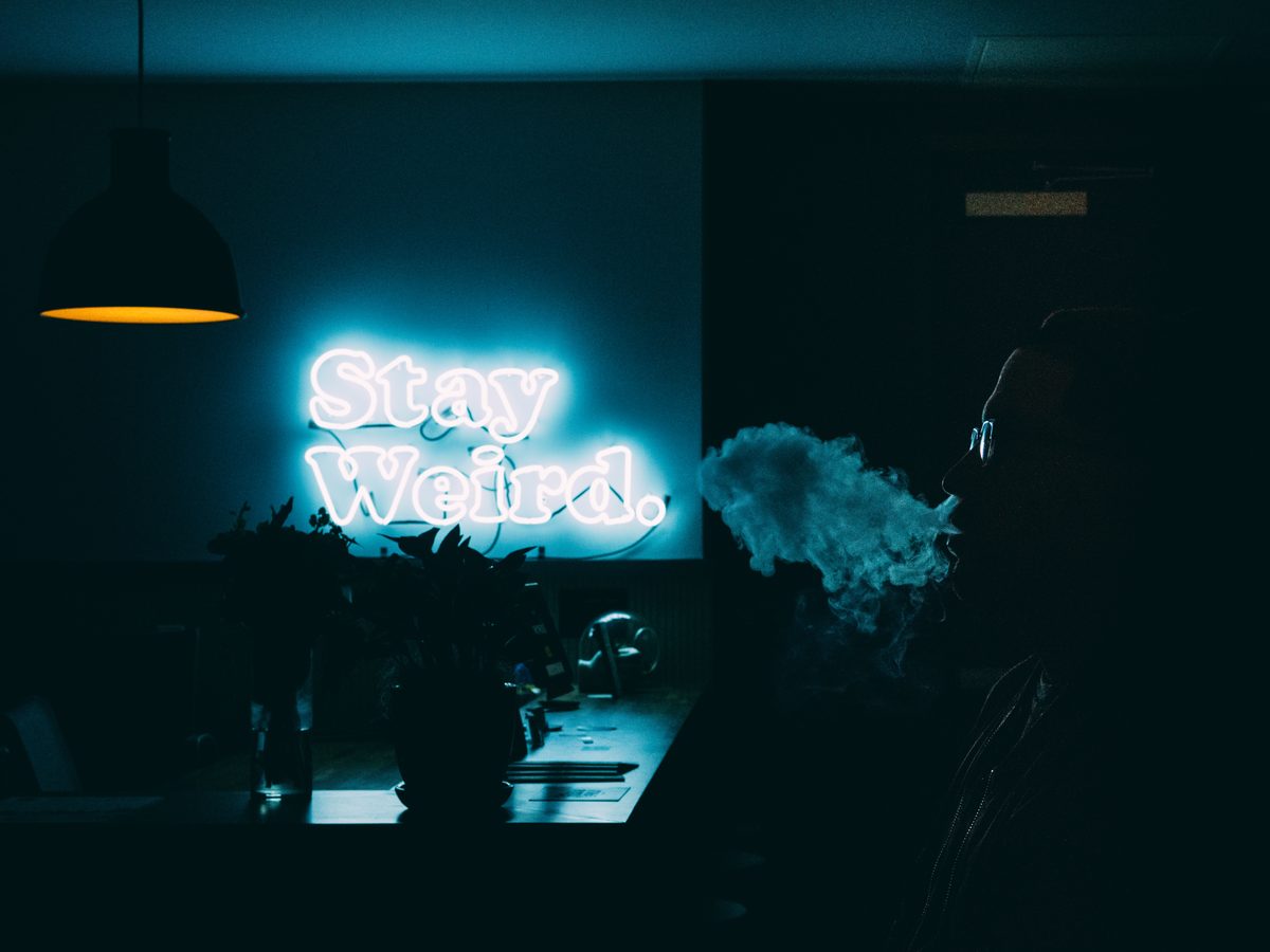 A figure exhaling vapour beside a Stay Weird neon sign in a dark room.