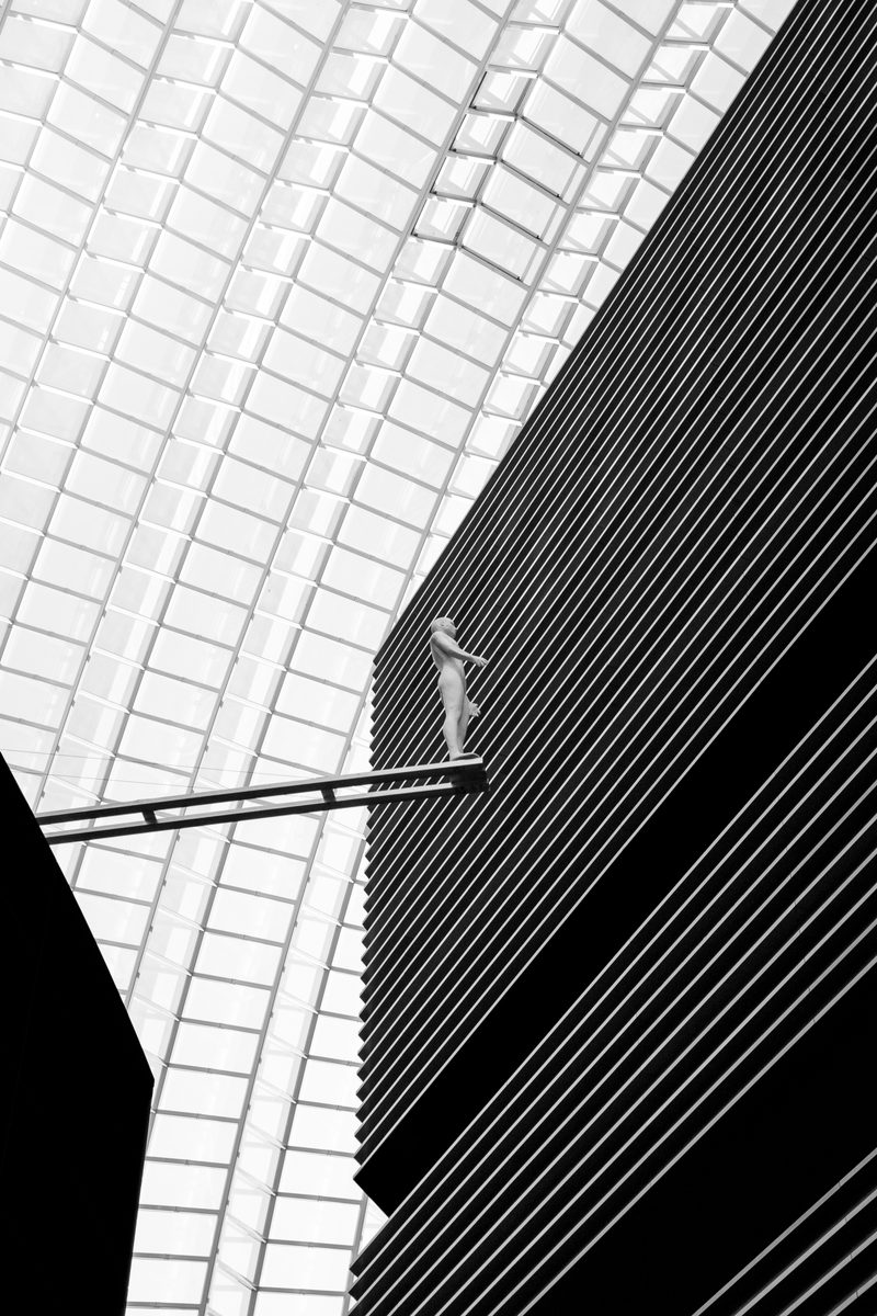 A lone figure on a walkway between a white glazed canopy and a dark ribbed facade, in black and white.