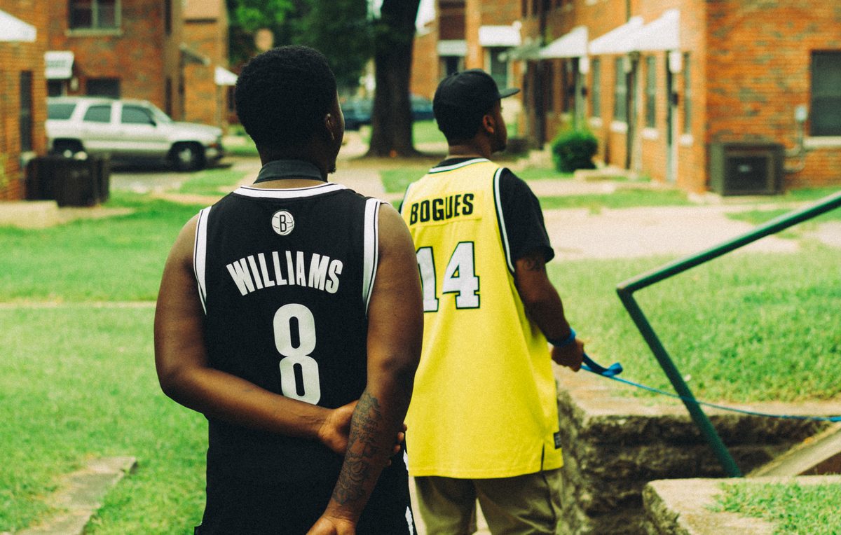Two men walking away between brick housing blocks in basketball jerseys, one carrying a hose.