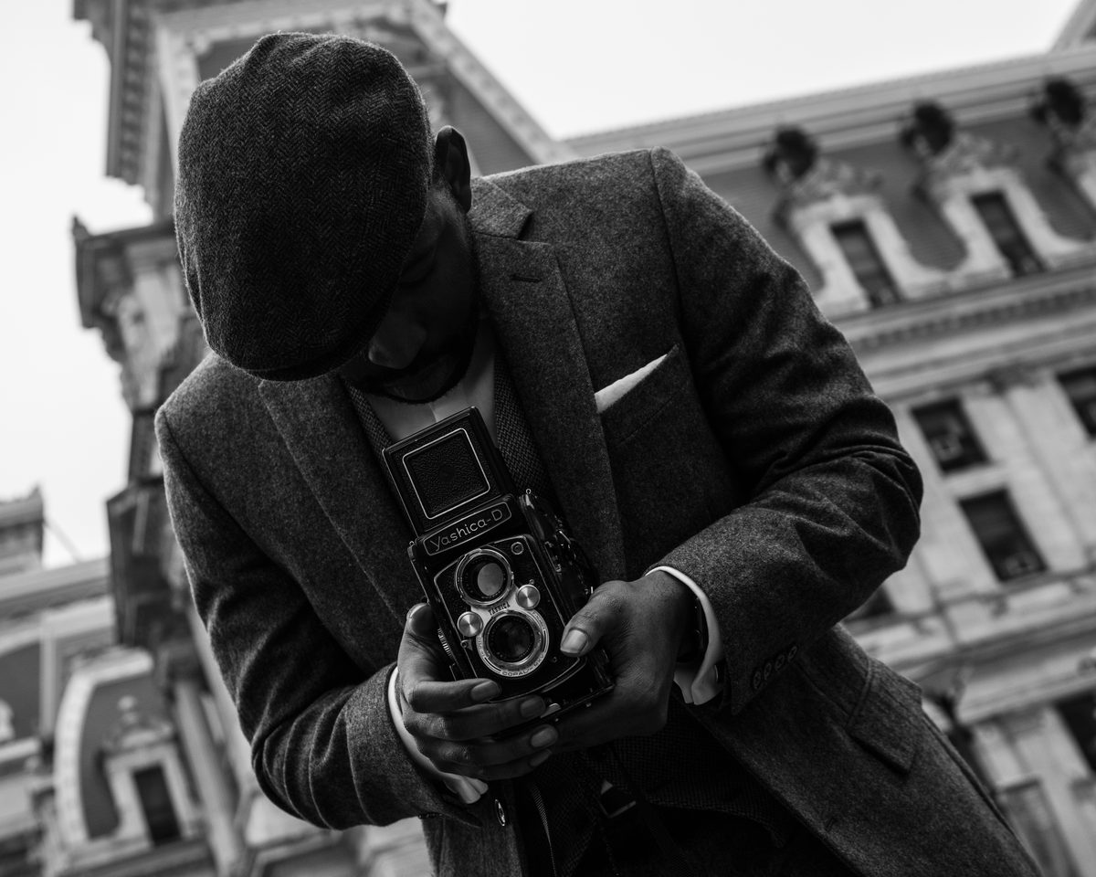 A man in a flat cap and tweed suit looking down into a twin-lens reflex camera, City Hall rising behind him.