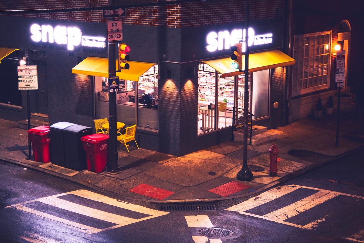 A lit corner shopfront with yellow awnings at night, empty crosswalks in front.