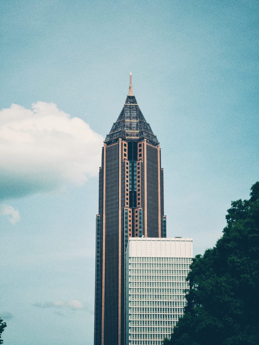 A tall tower with a latticed crown and spire rising past a lower white block into summer cloud.