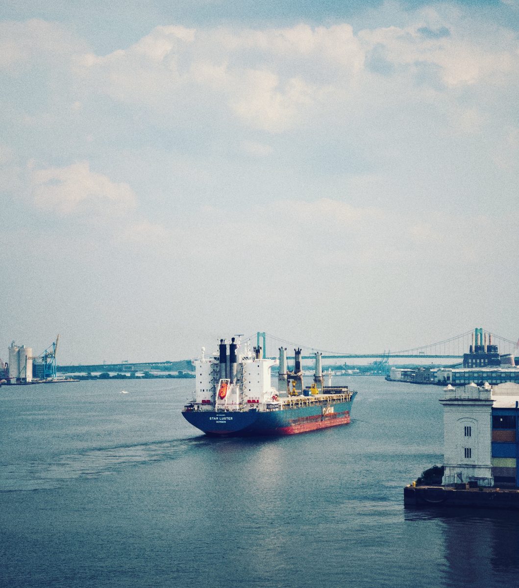 A cargo ship heading upriver past a pier, a suspension bridge and port cranes behind.
