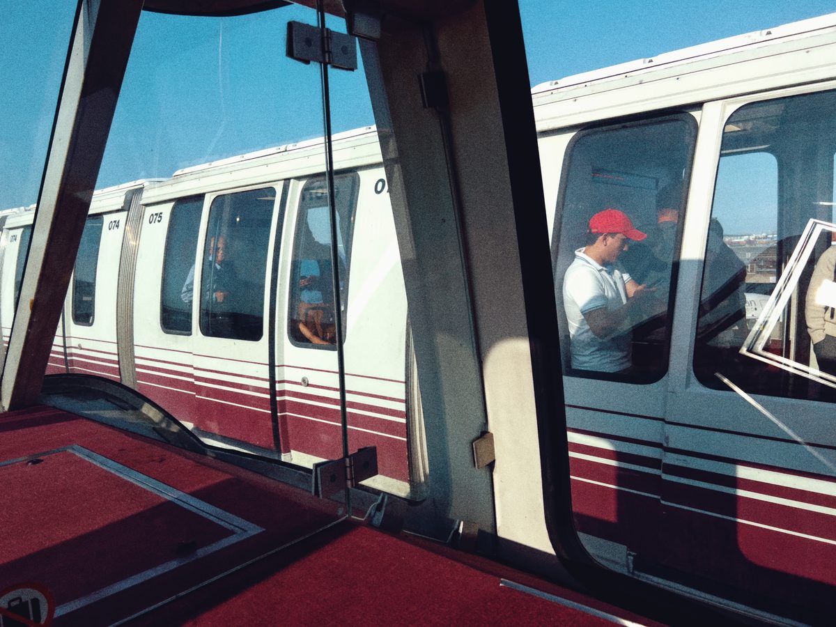 Passengers waiting inside an airport shuttle train, seen through its open doors in hard sun.
