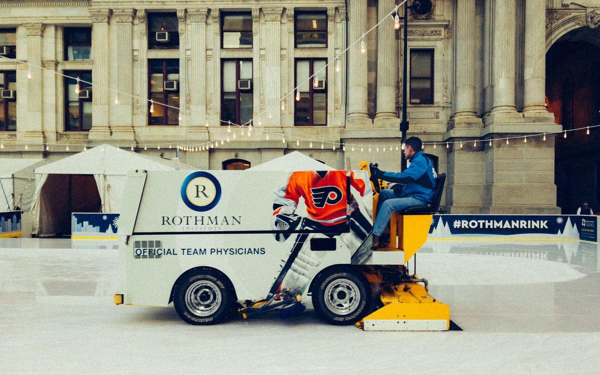 A Zamboni resurfacing the outdoor rink at City Hall, festoon lights strung overhead.