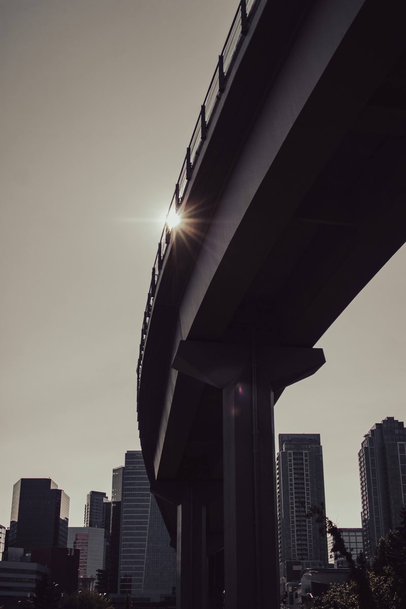 The underside of an elevated guideway curving away, the sun flaring off its edge above a downtown skyline.