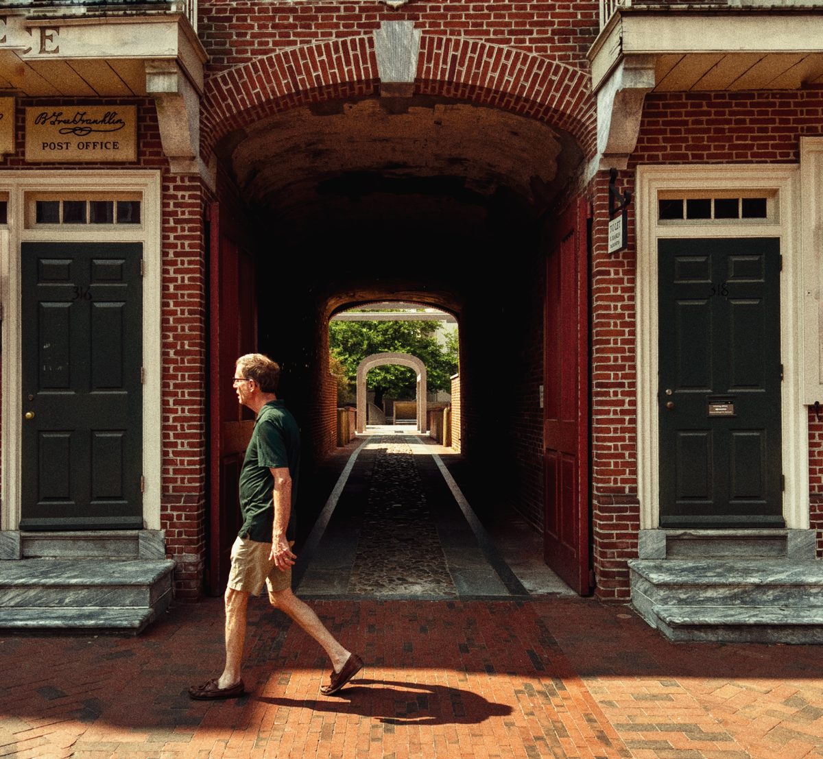 A man walking past the brick archway of an old post office, a run of arches receding into shadow behind him.