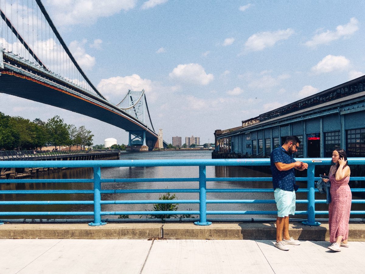 A couple on their phones at a blue waterfront railing, the Benjamin Franklin Bridge behind them in summer sun.