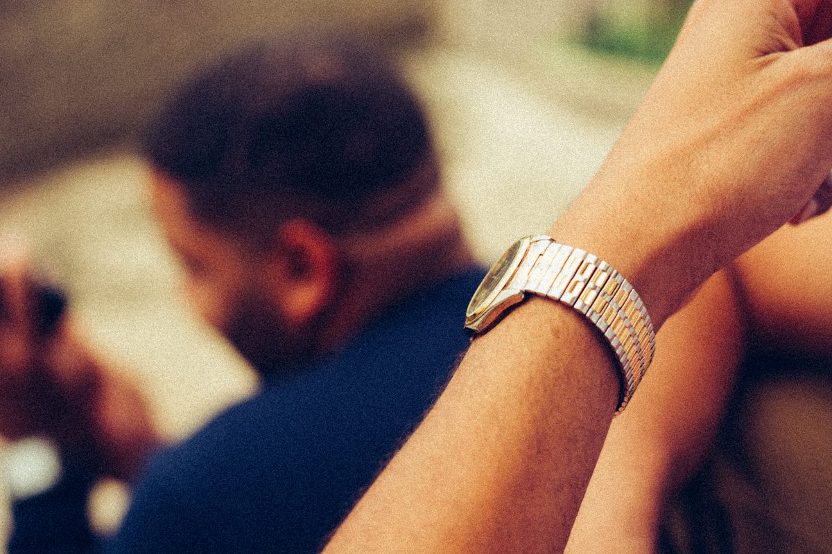 A gold bracelet watch on a wrist held up close, a figure out of focus behind.