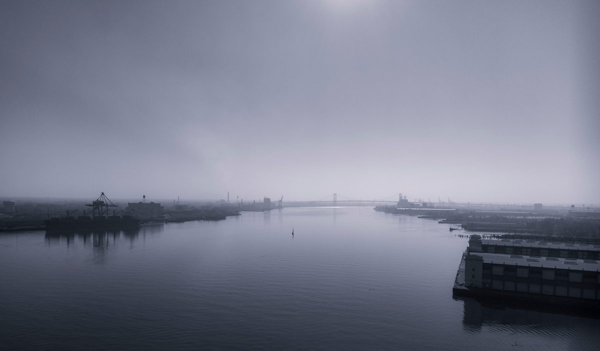 A river under thick fog, cranes and a distant bridge barely showing, the sun a disc overhead.