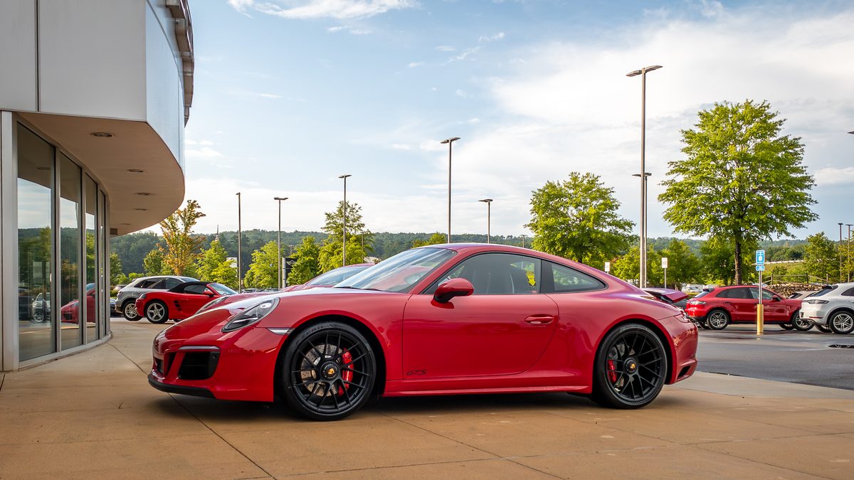 A red Porsche 911 GTS parked at a dealership entrance, wooded hills behind in summer light.