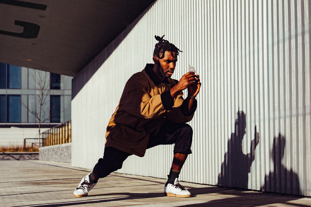 A man crouching against a corrugated white wall in hard sun, his shadow thrown sharp beside him.