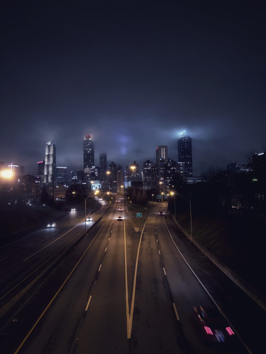 A highway running toward a skyline lost in low night cloud, tail-lights trailing at the frame edge.