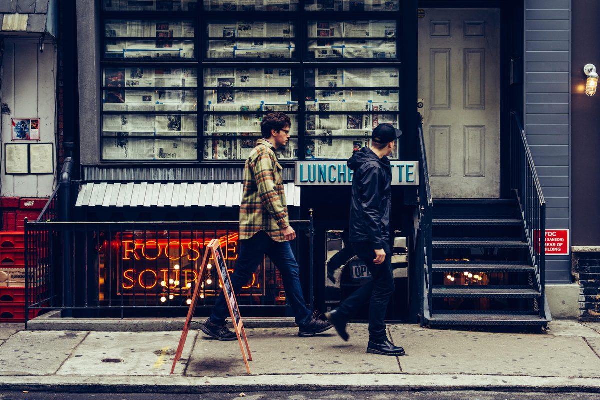 Two men passing a luncheonette storefront stacked with newspapers, its neon lit behind the railing.