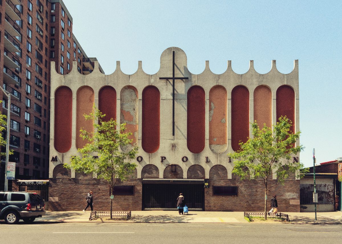 The arched concrete front of the Metropolitan church, red panels weathered, pedestrians passing in summer.