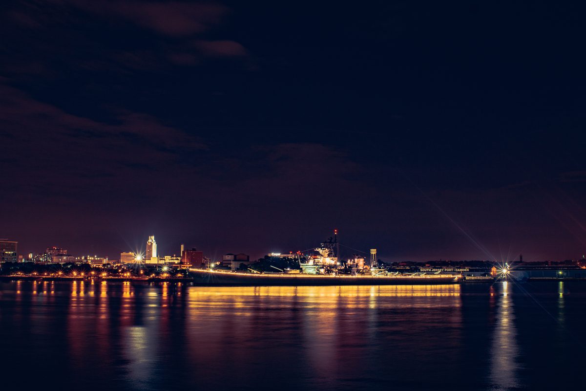 A battleship moored across the river at night, its lights drawn out in long reflections on the water.