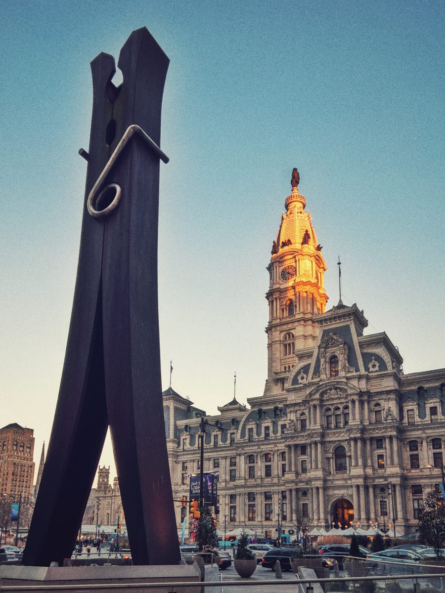 Claes Oldenburg's Clothespin standing against the lit tower of City Hall at dusk.