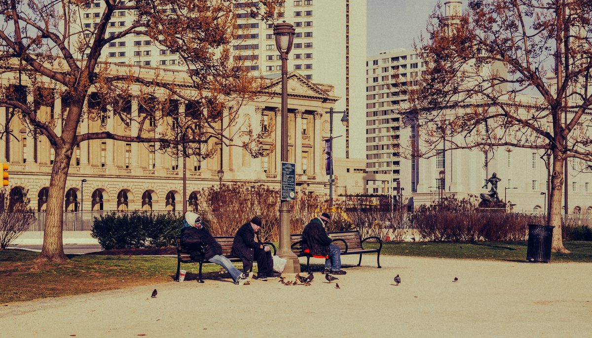 Three people on benches in the park by City Hall in winter, pigeons on the gravel at their feet.