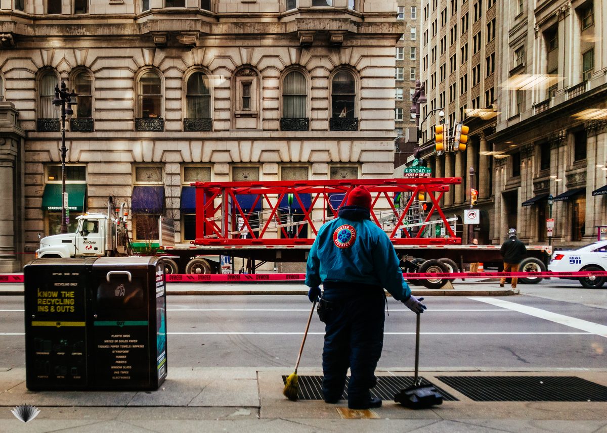 A street cleaner watching a red steel truss go by on a flatbed, on Broad Street in winter.