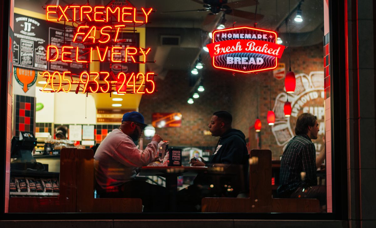 Diners at a table behind a restaurant window, neon advertising fast delivery across the glass.