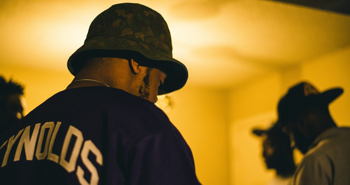 A man in a camouflage bucket hat seen from behind in a warm-lit room, a second figure out of focus beyond.