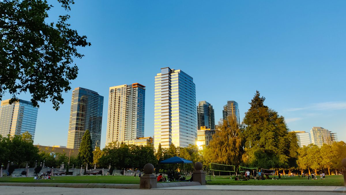 A downtown park at golden hour, glass towers over the trees and people out on the grass.