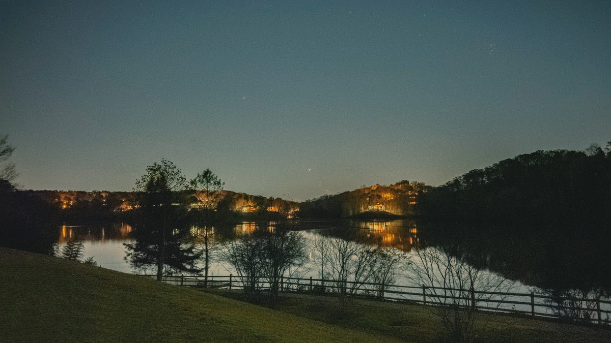 A reservoir at dusk, lights on the far shore doubled in still water below wooded hills.