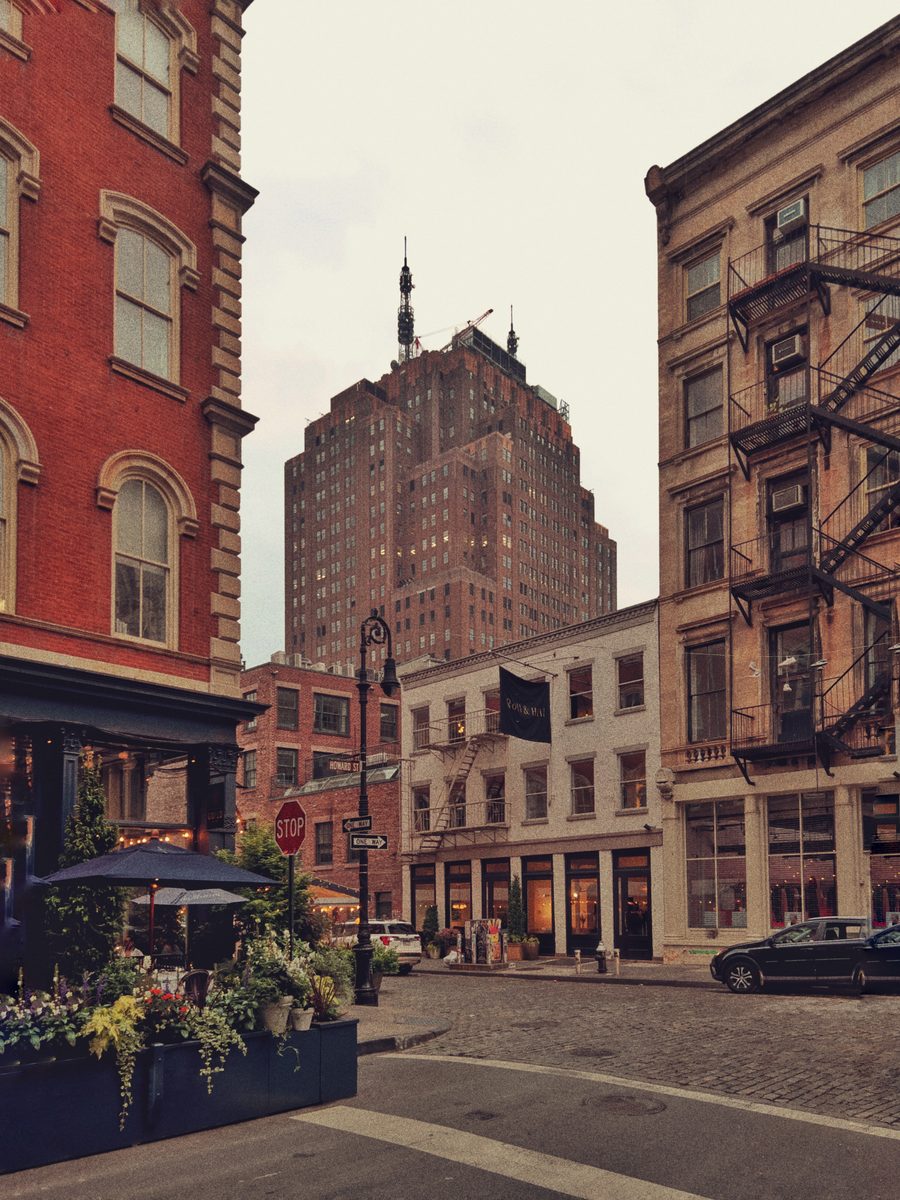 A cobbled Tribeca corner in soft light, cast-iron fronts and fire escapes under a setback tower, flowers outside a cafe.